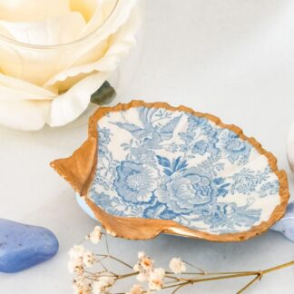 Scallop shell jewellery dish decorated with a blue and white toile design and gilded in gold. The dish rests on a white surface surrounded by blue lace agate and white flowers.