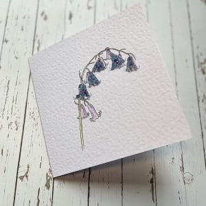 A greetings card lays on a table, features a single stem bluebell flower with watercolour and fabric flower detail