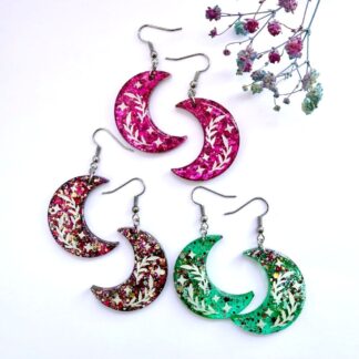Three pairs of handmade glitter crescent moon resin earrings in pink aurora, hot pink and teal by Copper Magpie Designs UK. Earrings are hung on surgical steel ear hooks. Displayed on a white background with blue and purple flowers.