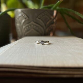 a dainty silver ring, all polished with a small lavender coloured stone set into a bezel is facing straight at the camera. It is laying on an open book.