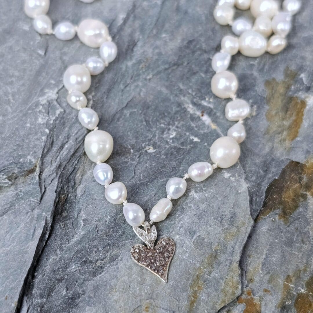 White baroque pearl necklace with a small etched silver heart pendant on a silk knotted strand, flat rock background, Isobel Kelly Jewellery