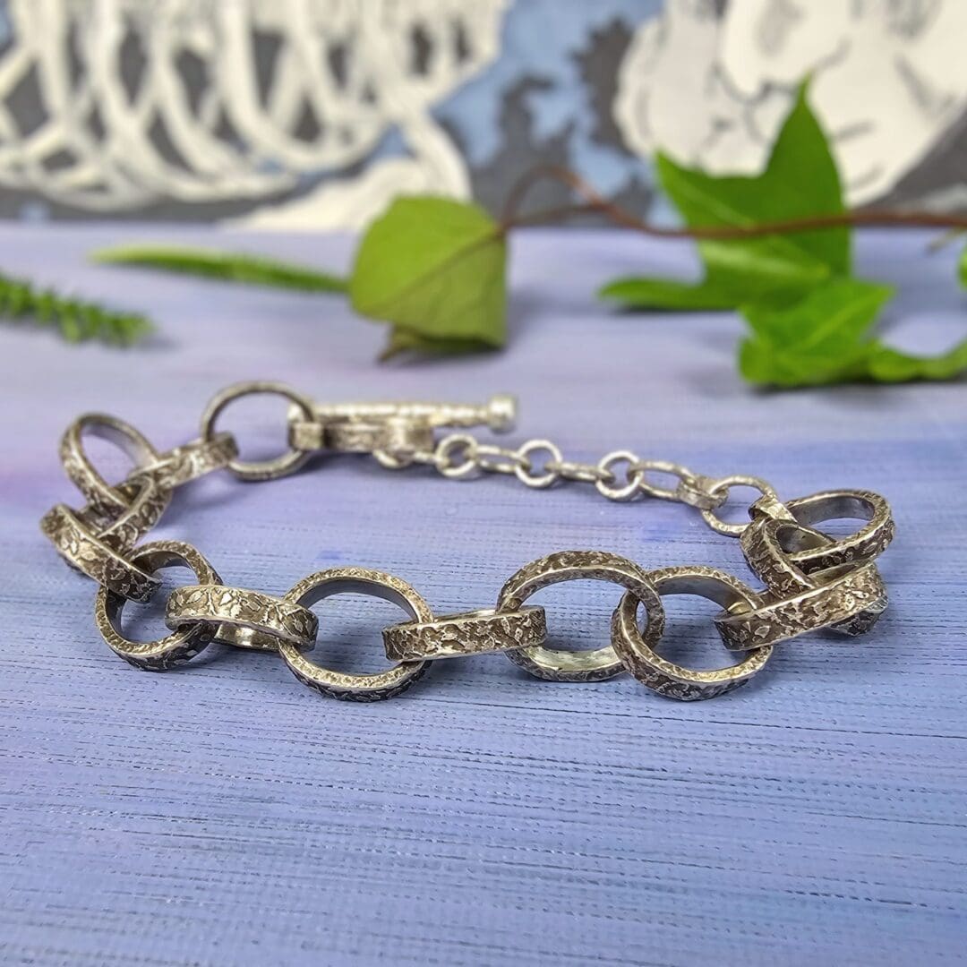 Chunky silver chain bracelet with hand etched leafy patterns and a forged toggle clasp, antique finish, lilac background, Isobel Kelly Jewellery