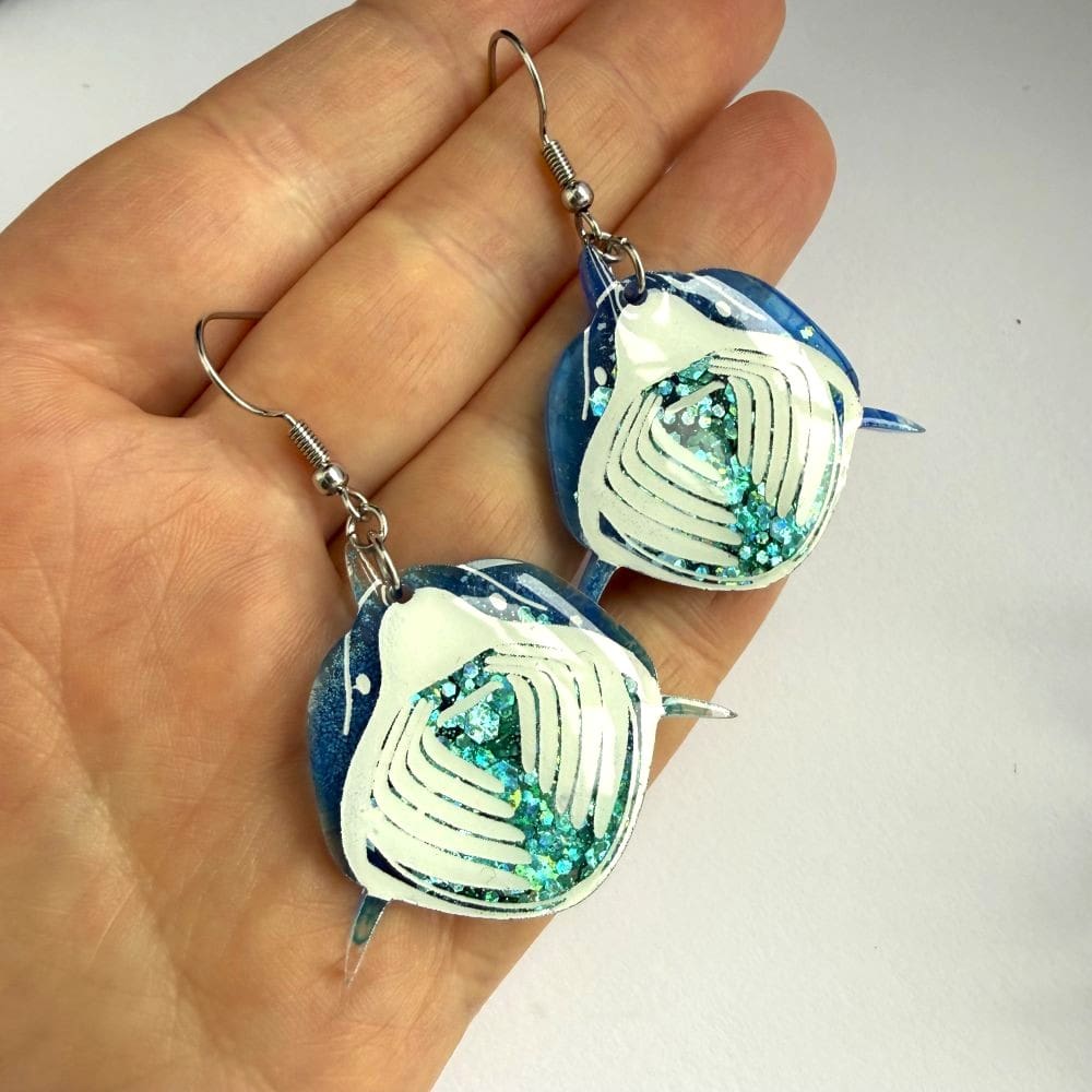 Close up of handmade ocean blue mist basking shark earrings with blue/green holographic glitter displayed on a hand for scale. 50p from the sale of each pair of earrings goes to marine charities.