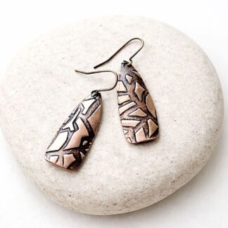 A pair of copper textured drop elongated earrings with copper ear wires. Displayed on a white surface resting on a stone.