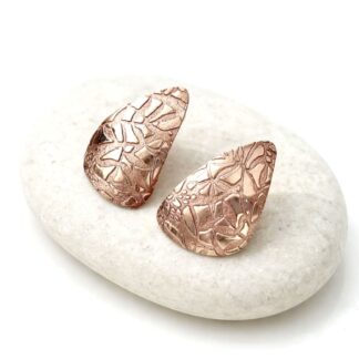 A pair of large handcrafted textured copper stud earrings. They are displayed sat on a white surface resting on a stone.