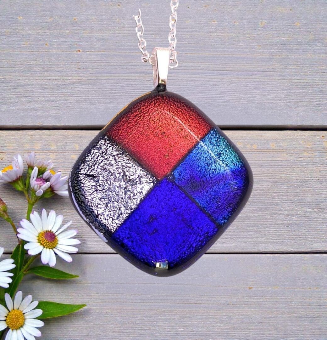 Square pendant necklace with red, blue, purple, and silver textured glass sections on a silver chain, next to white daisies on gray wood.