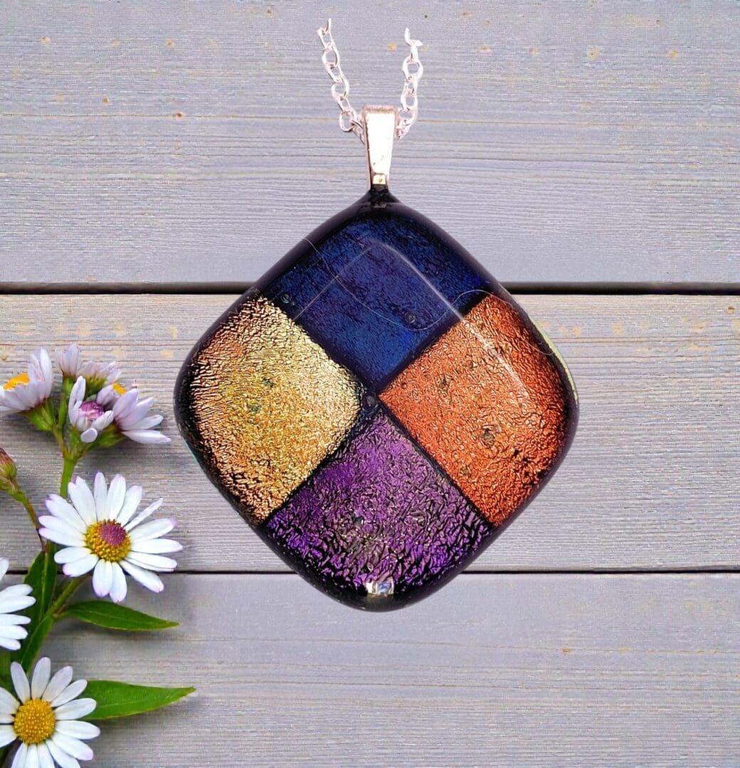 Square glass pendant necklace with metallic gold, blue, orange, and purple sections, placed on wooden surface next to white daisies.