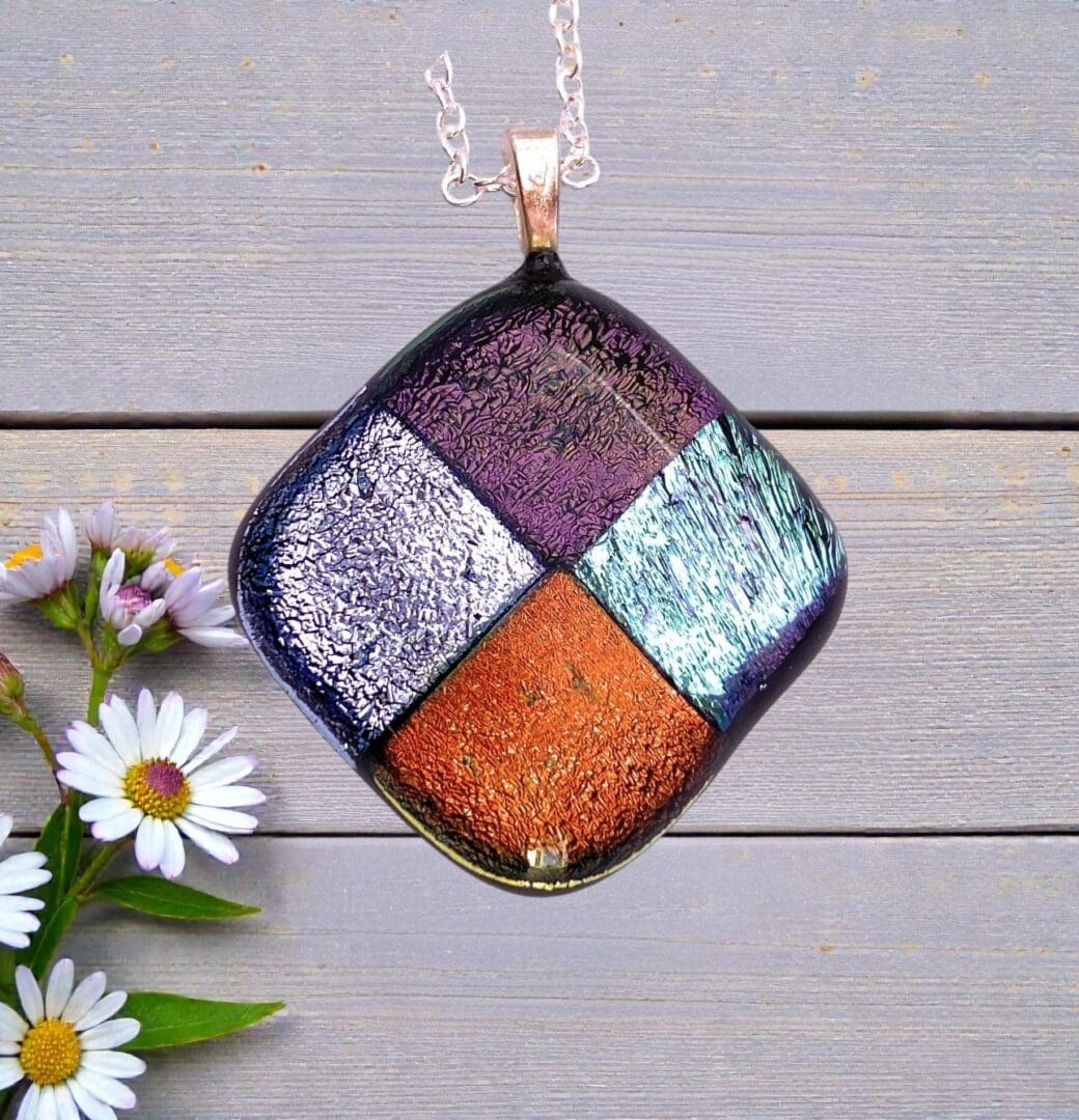 Square glass pendant with purple, silver, turquoise, and copper color blocks on a silver chain, displayed on wooden planks with daisies.