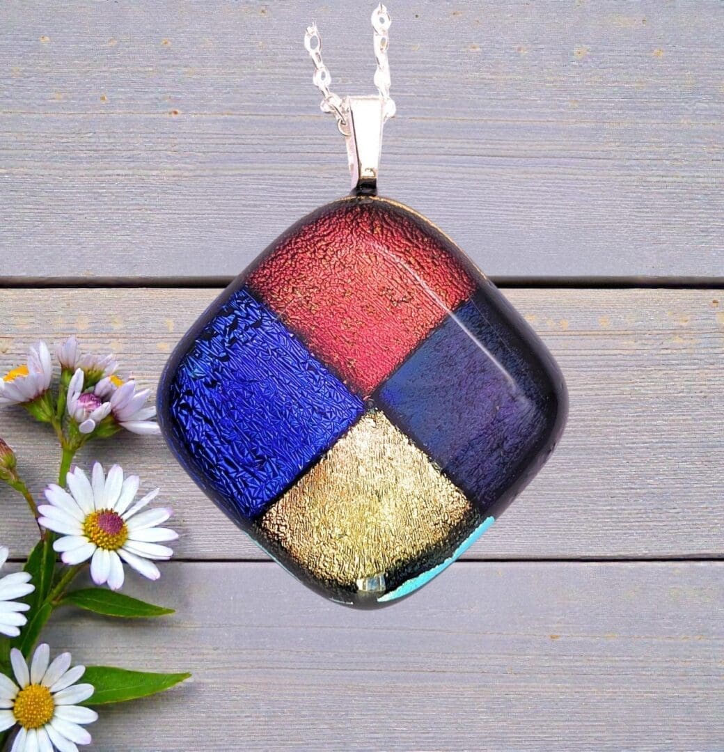 Square glass pendant with red, blue, purple, and gold textured sections on a silver chain, resting on wooden surface with daisies.