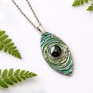 A handcrafted fine silver almond shaped pendant that is textured and hand painted in metallic blue and green paint and has a sterling silver chain. It is displyed on a white background leaning against a white background.