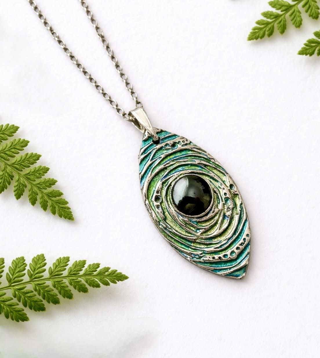 A handcrafted fine silver almond shaped pendant that is textured and hand painted in metallic blue and green paint and has a sterling silver chain. It is displyed on a white background leaning against a white background.