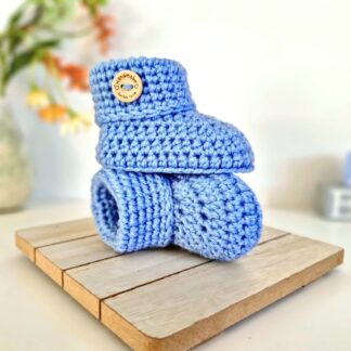 a pair of handmade crochet baby booties in blue featuring a folded cuff and a round wooden button on a neutral background.