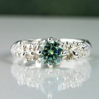 Handmade sterling silver ring with a textured organic band, set with a round green moissanite gemstone, photographed in soft natural light with a subtle reflection.