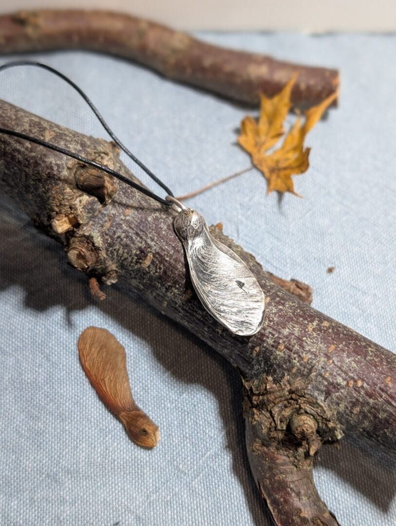 A silver sycamore seed hanging from a leather cord displayed on a small branch with an autumn leaf and a real sycamore seed