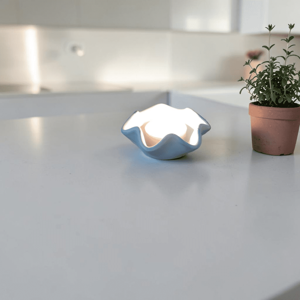 A small tea light holder with a lit candle in it sits on a white kitchen counter next to a plant pot with pink flowers in.
