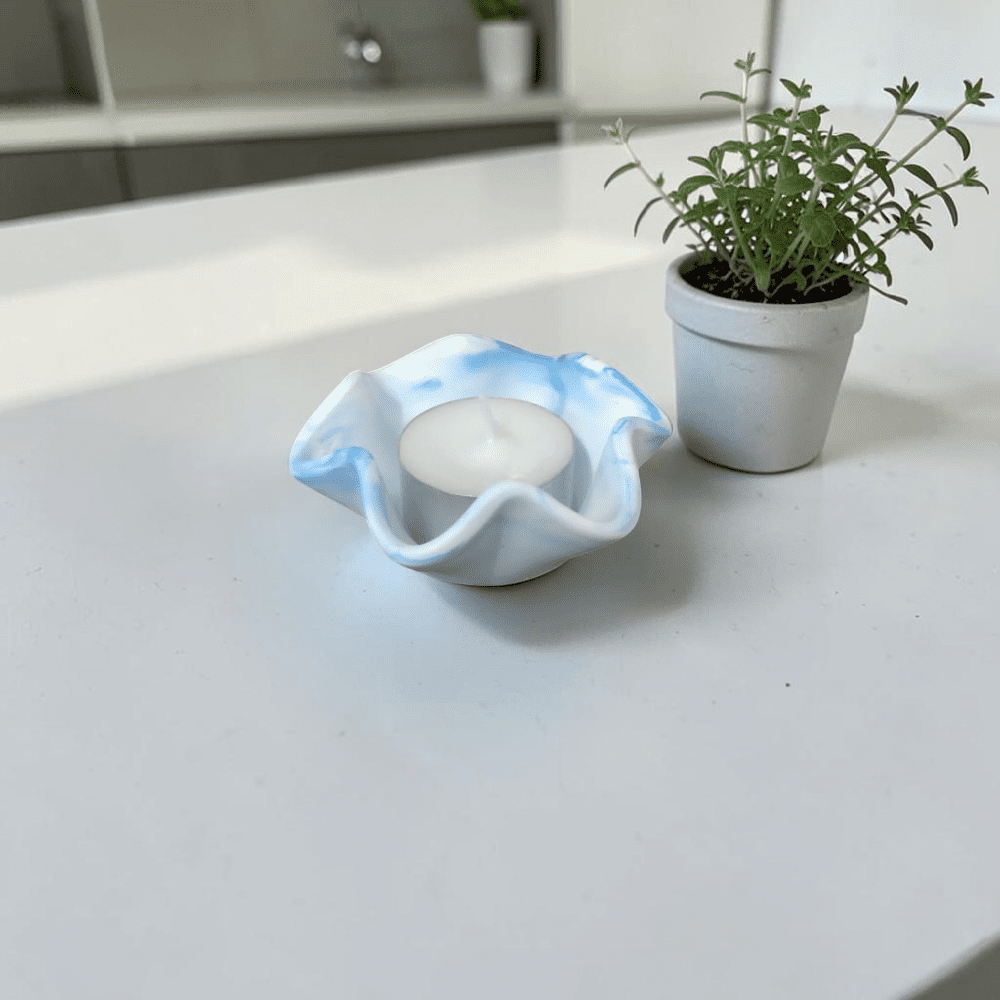 A small blue marble patterned teal light holder with a wavy lip, sits on a white countertop next to a small, white plant pot with a green plant in it.