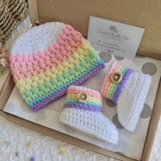 A matching handmade baby gift set featuring a white crochet beanie and booties, both with soft pastel rainbow stripes.