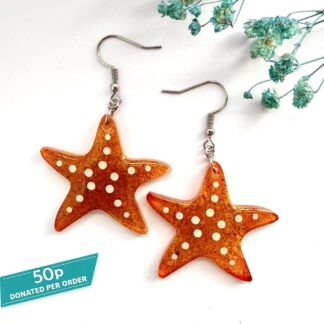A pair of orange starfish or sea star resin earrings containing sand from Formby beach in Merseyside made by Copper Magpie Designs, UK. Displayed on a white background with blue flowers.