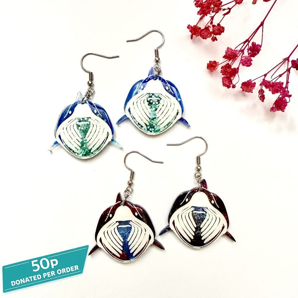 Picture shows two variations of handmade basking shark resin earrings, one in ocean blue mist with green/blue glitter gill detail and one in black night with red tint glitter and grey gill detail by Copper Magpie Designs UK. 50 p donation to marine charities for each order (donated annually).