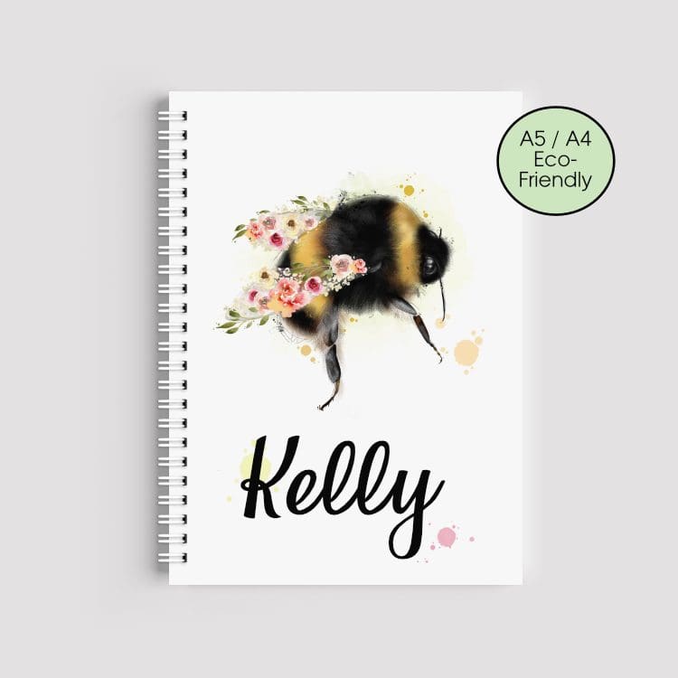 A spiral-bound A5 notebook featuring a watercolor illustration of a large bumblebee with pink and peach flowers for wings. The name "Kelly" is printed in black script at the bottom. An "Eco-Friendly" badge is in the corner against a clean grey background.