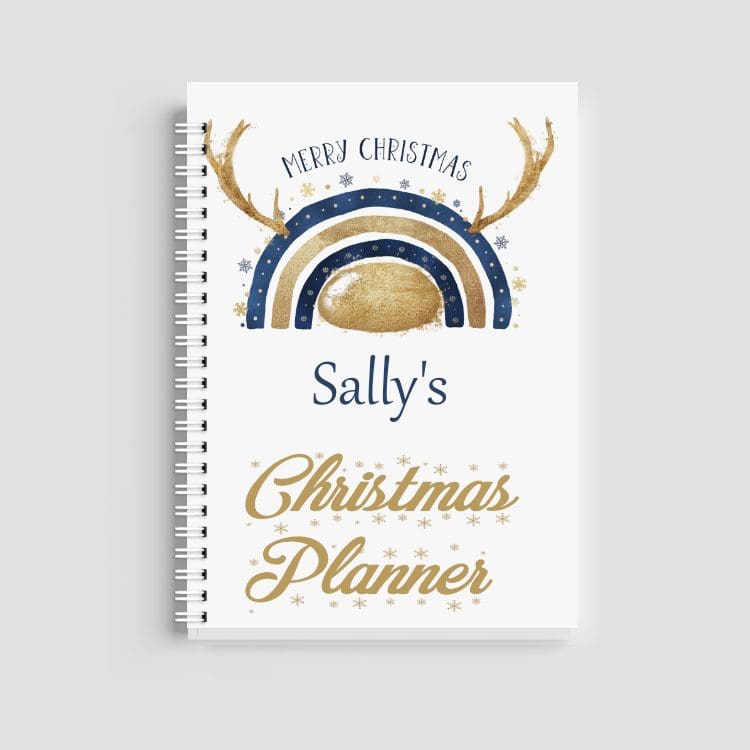 A spiral-bound notebook with a white cover. It features a navy and gold rainbow with reindeer antlers at the top, the name "Sally's" in navy, and "Christmas Planner" in gold script at the bottom.