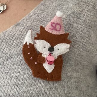 Handmade fox birthday badge, the fox is wearing a party hat and is holding a pink cupcake