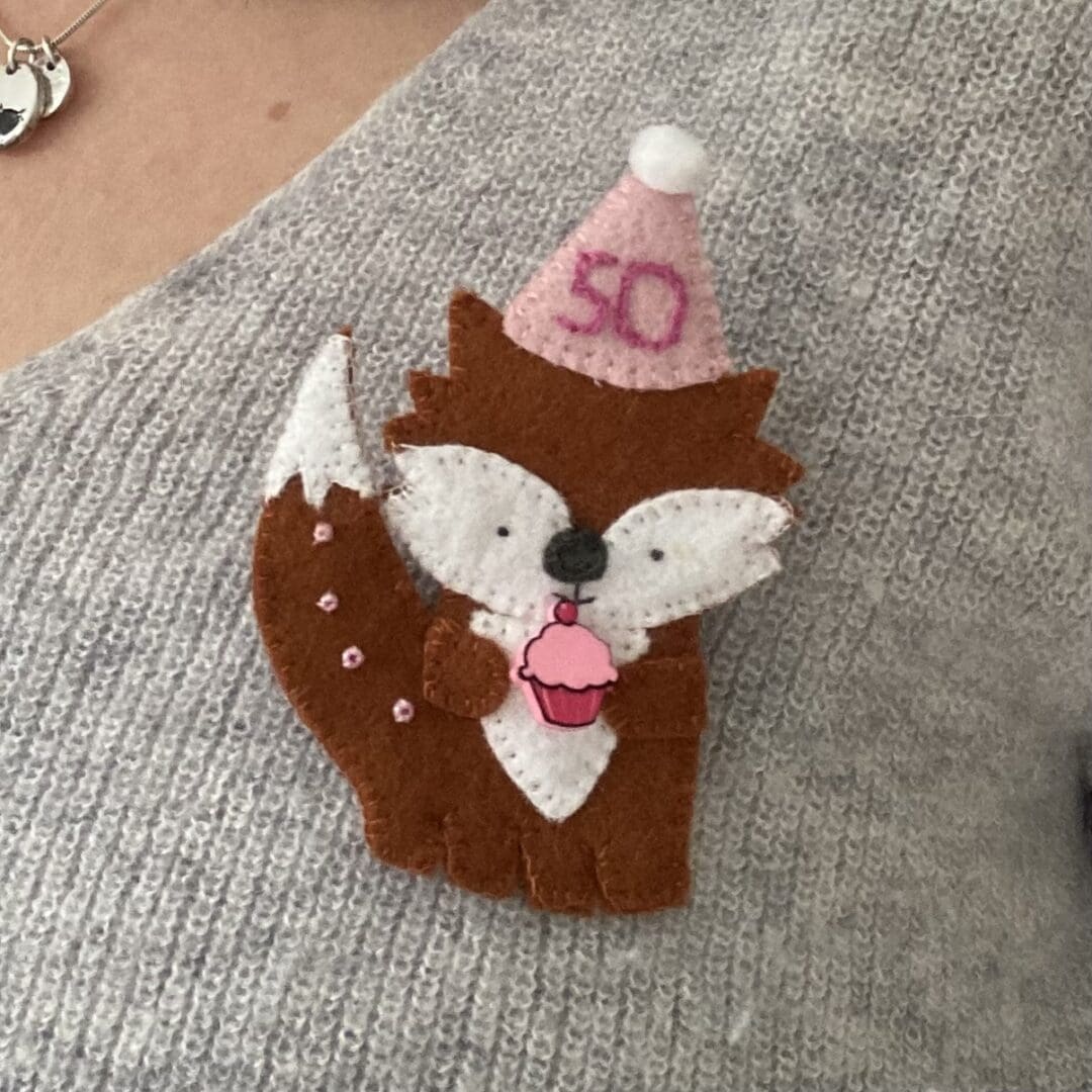 Handmade fox birthday badge, the fox is wearing a party hat and is holding a pink cupcake