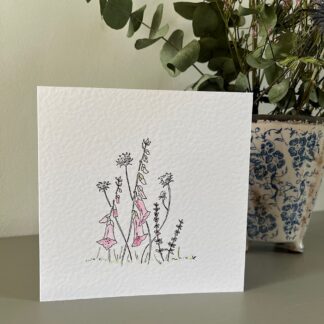 A card with hand drawn and painted cottage garden flowers in pinks and purples. Stands in front of a pot of flowers.