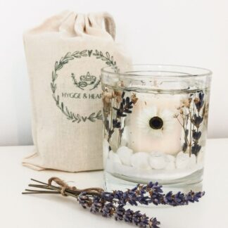 Daisy and lavender refillable gel and soy wax candle with real flower and shell embeds on a white surface next to sprigs of lavender and a cotton drawstring bag.