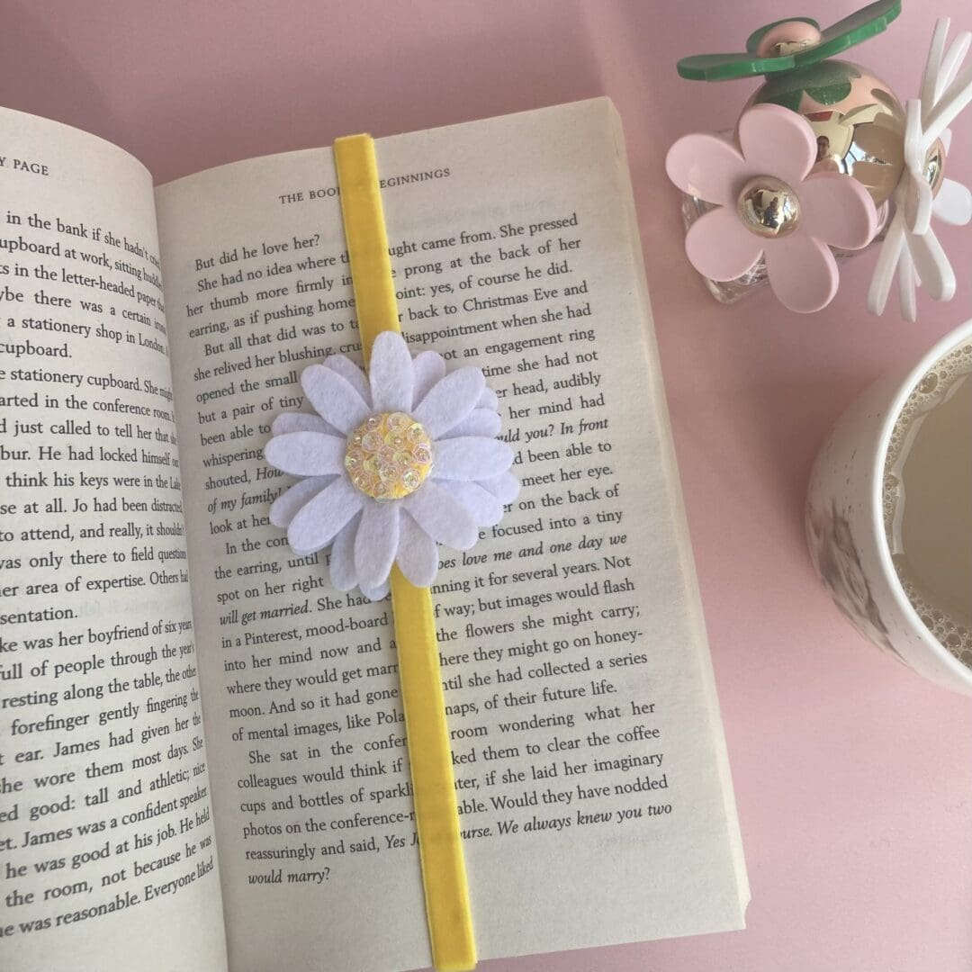 White felt daisy with a yellow centre, the centre is filled with sequins that shimmer in the light with a bright yellow velvet ribbon holding a page in a book