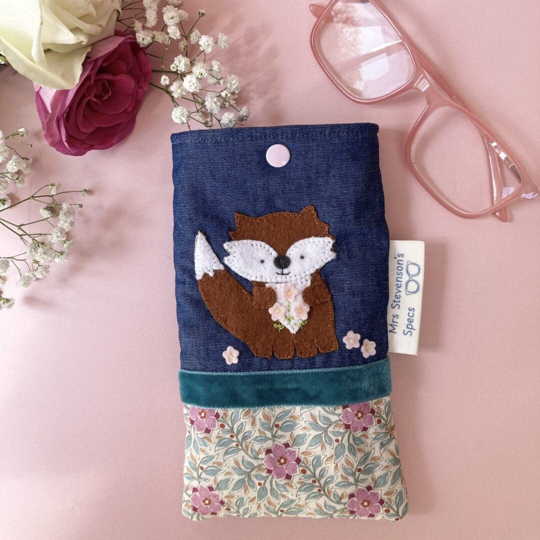 Handmade glasses case top section is denim which has felt fox sewn on it. The fox has a white face and tummy. The fox is holding 3 pretty pink felt flowers. The lower section is a pretty cream fabric with pink flowers. I had separated the denim and pretty fabric wit h a teal velvet ribbon. The glasses case closes with a pale pink snap fastener.