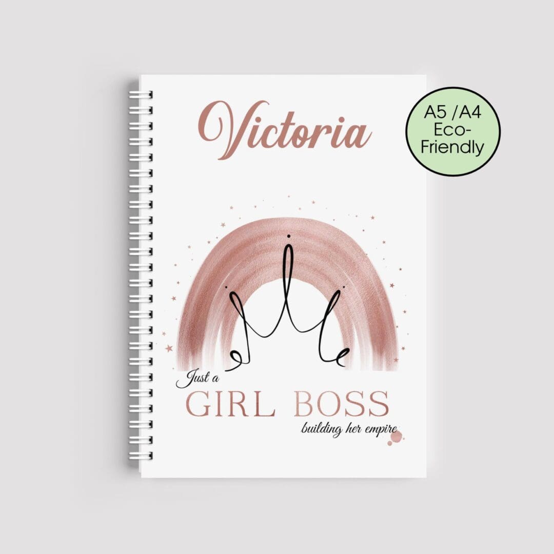 A spiral-bound notebook with a white cover. It features the name "Victoria" in rose gold, a watercolor rainbow with a black crown outline, and the text "Just a GIRL BOSS building her empire" at the bottom.