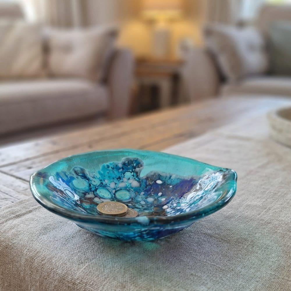 Sculptural turquoise and cobalt blue fused glass trinket bowl with wave-like edges, displayed on a wooden coffee table in a bright living room.