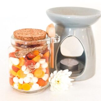 Glass jar of soy wax scoopies in white, yellow and orange with a cork lid and wooden scoop stands on a white background next to a grey wax burner and white flower.