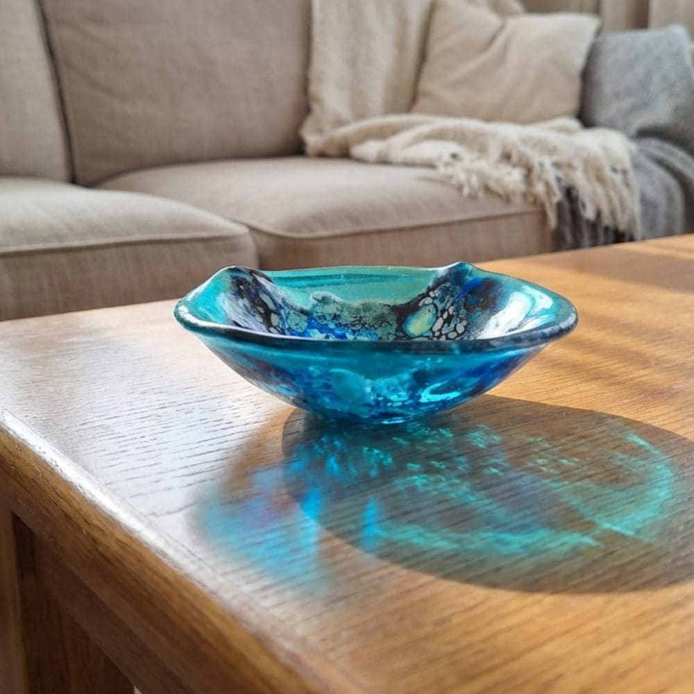Organic shaped turquoise fused glass trinket bowl with vibrant blue frit lace pattern, displayed on a wooden coffee table in a living room.