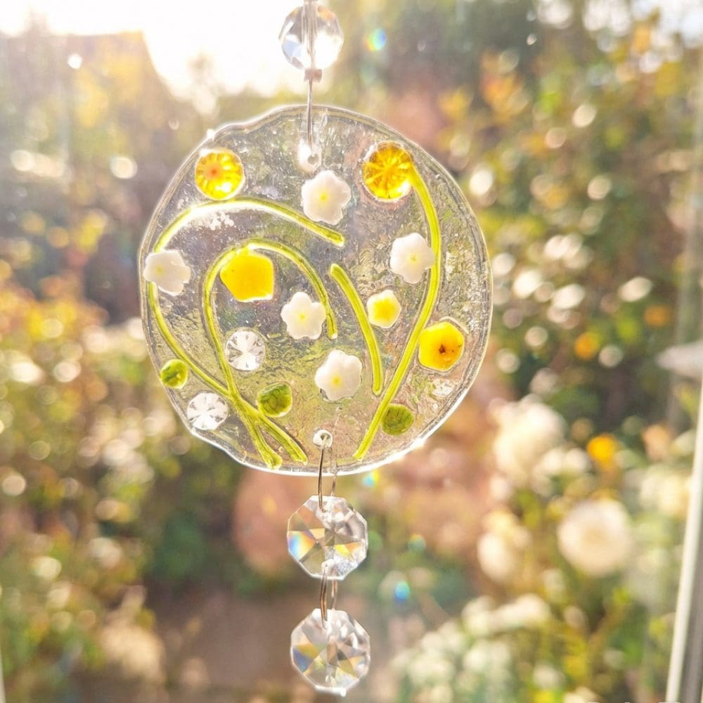 Fused glass floral suncatcher catching sunlight in a window, showing yellow and white meadow flowers