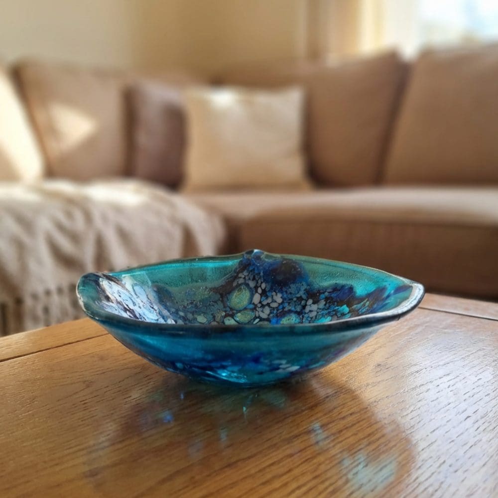 Side view of a handmade turquoise and blue frit lace glass trinket bowl resting on a wooden coffee table with a sofa in the background