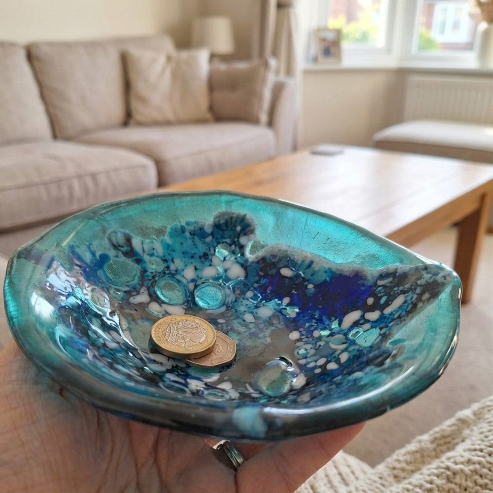 Close-up of a kiln-fired fused glass trinket dish in shades of turquoise, cobalt blue and iridescent colours, placed on a wooden table.