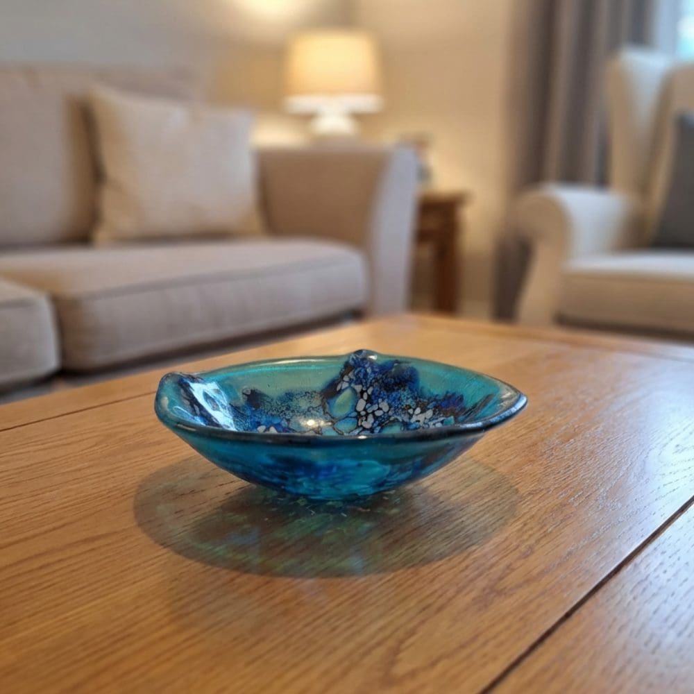 Turquoise fused glass trinket bowl displayed on a wooden table in a living room, featuring cobalt blue and iridescent frit lace pattern.