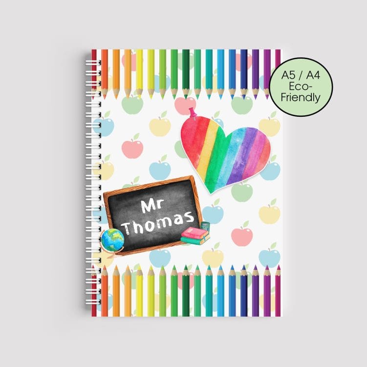 A spiral-bound A5 notebook with a rainbow pencil border at the top and bottom. The background features a pattern of colorful apples, a rainbow heart, and a chalkboard with the name "Mr Thomas" written on it.