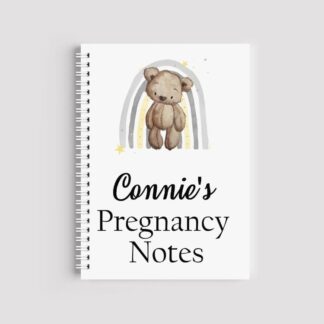 A spiral-bound A5 notebook with a white cover featuring a watercolor teddy bear in front of a grey and yellow rainbow. The text reads "Connie's Pregnancy Notes" in a mix of script and serif fonts.