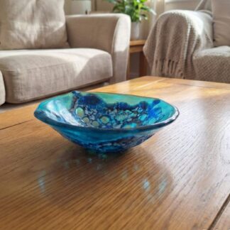 Handmade turquoise frit lace fused glass trinket bowl on a wooden coffee table, showing iridescent blue and turquoise colours with organic wavy edges.