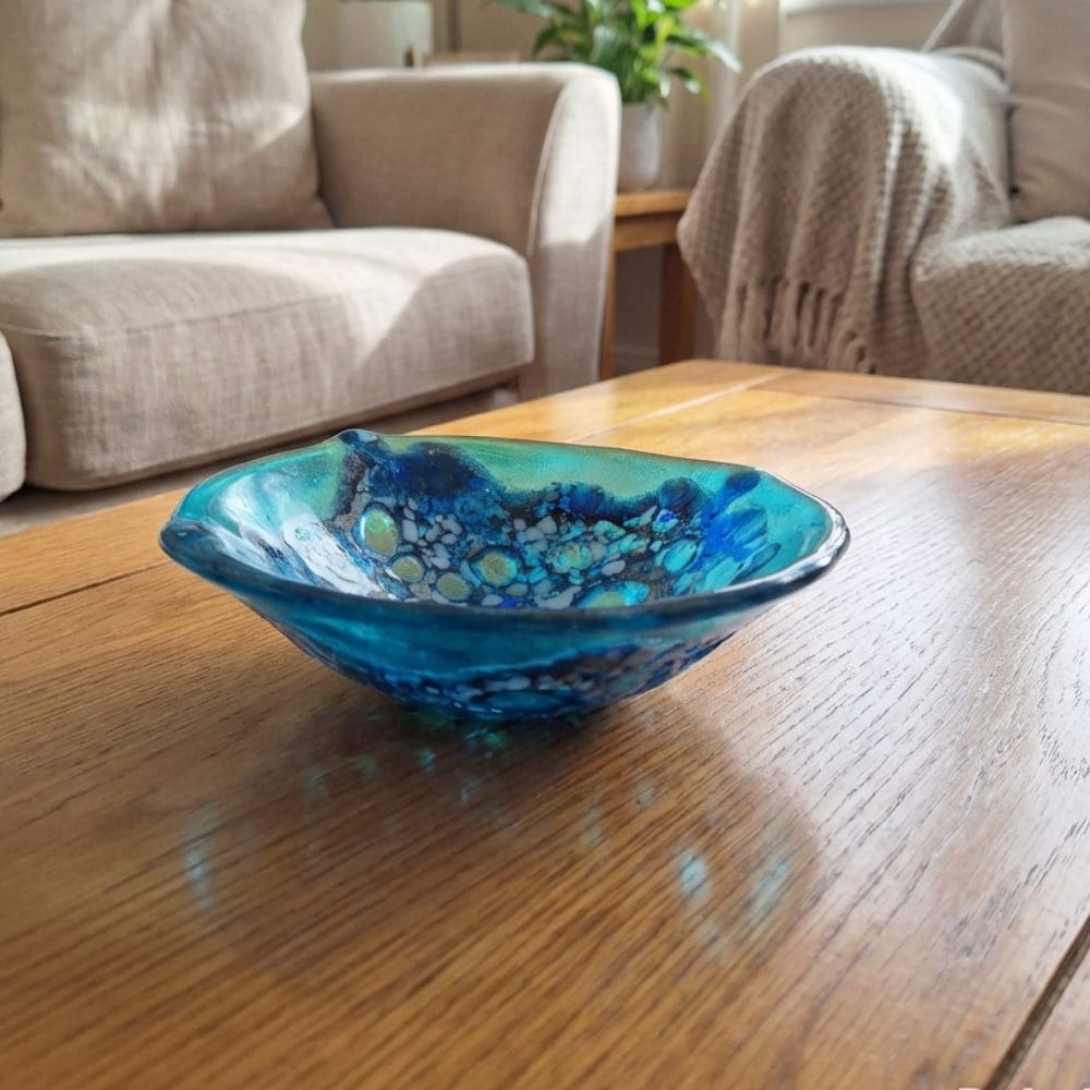 Handmade turquoise frit lace fused glass trinket bowl on a wooden coffee table, showing iridescent blue and turquoise colours with organic wavy edges.