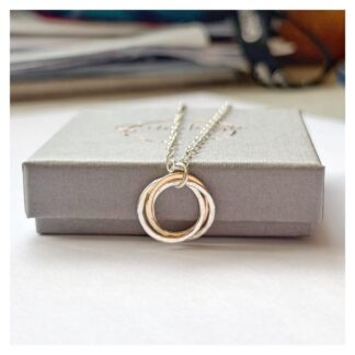 Pendant made from two silver rings and one gold ring entwined lies on a grey gift box. The pendant is attached to a silver cable chain.