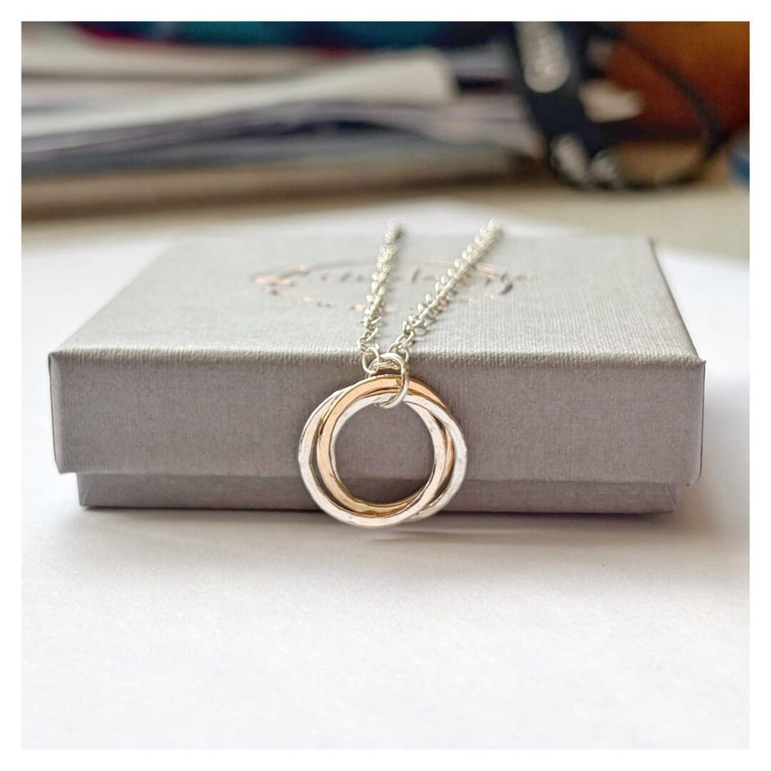 Pendant made from two silver rings and one gold ring entwined lies on a grey gift box. The pendant is attached to a silver cable chain.