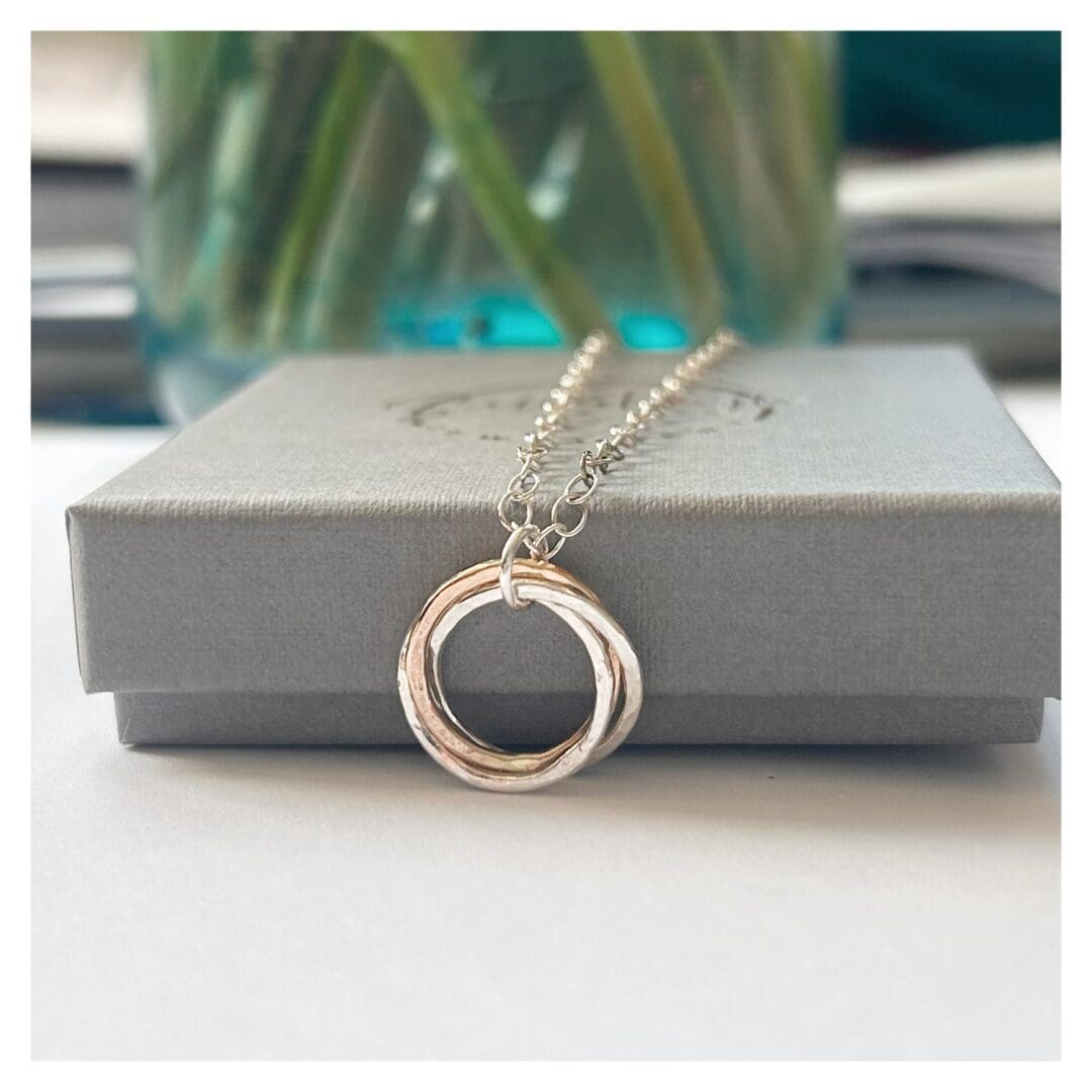 Pendant made from two silver rings and one gold ring entwined lies on a grey gift box. The pendant is attached to a silver cable chain.