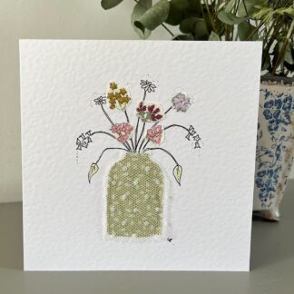 Greetings card with an appliqued vase with fabric and hand drawn dainty flowers cascading out of the vase. Stands in front of a pot of flowers