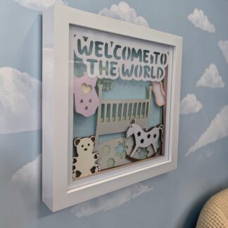 A layered paper art work for a new baby. It features a crib above a rug with a flower design. A rocking horse, bottle, onesie and teddy. All layered to make a 3D image. It is in a white box frame.