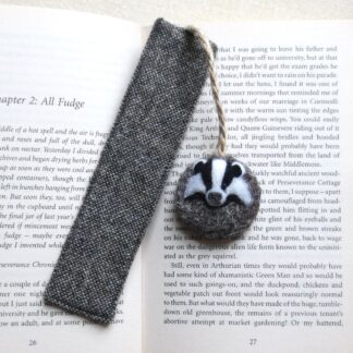 Grey flecked wool fabric bookmark laid across an open book. Attached to the top with a length of jute string is a wool disc with a badger face on it.