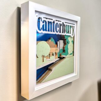 A layered paper art picture of Canterbury in Kent looking from its left side. Seen from Westgate gardens along the river Stour towards Westgate towers. The background is blue mirror card with the name Canterbury at the top of the front layer. It is in a white box frame.